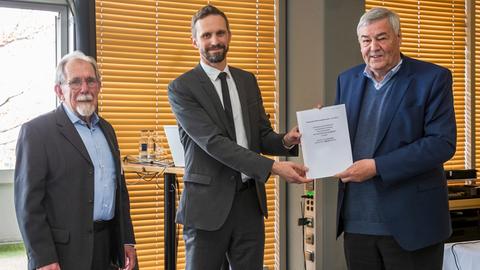 Innenaufnahme zeigt drei Männer nebeneinander stehend bei der Überreichung eines Dokuments